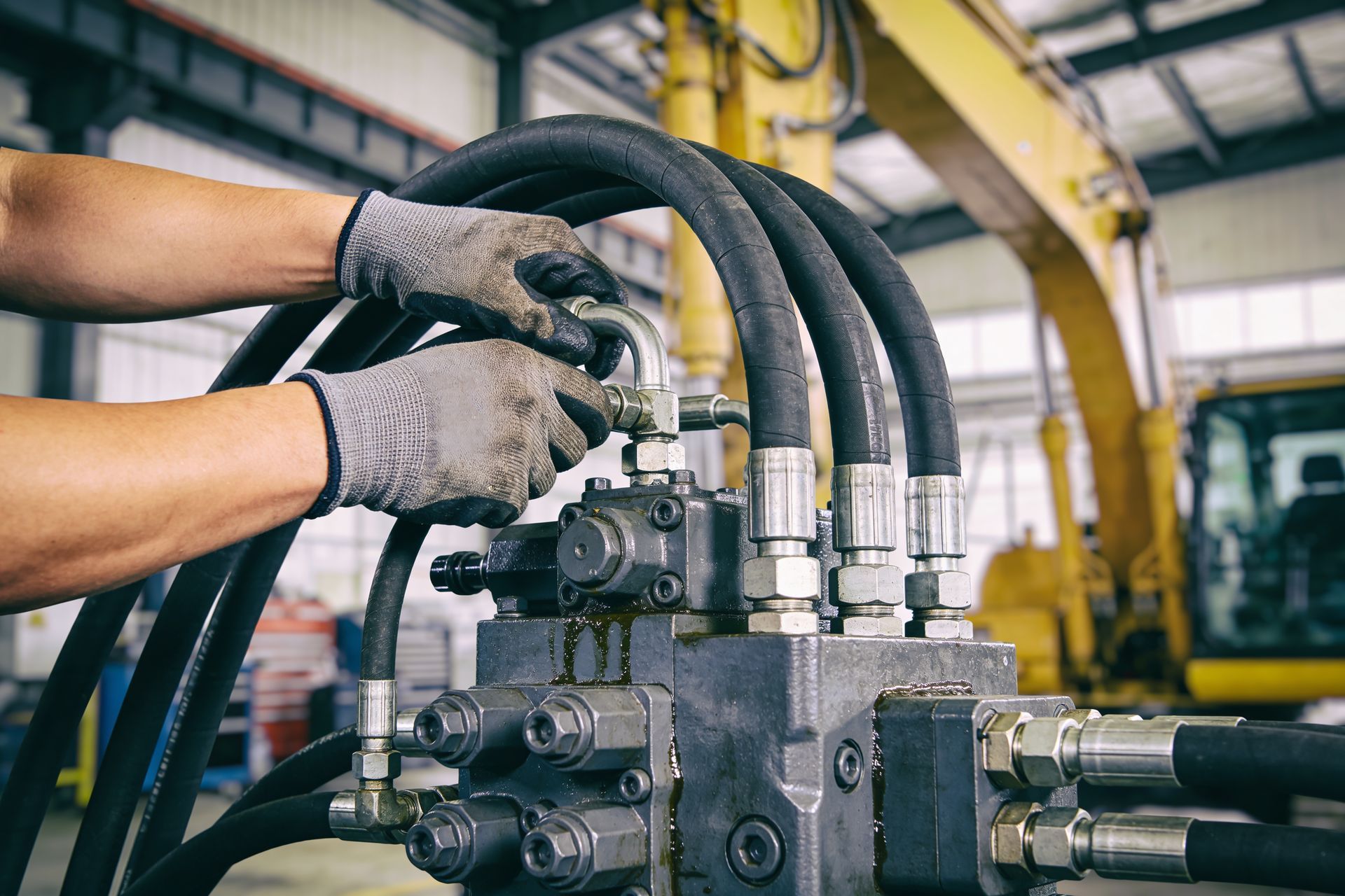 A mechanic uses a yellow and black hydraulic equipment from a supplier. A mechanic's hands in gloves are connecting hydraulic hoses to a heavy machinery valve block.