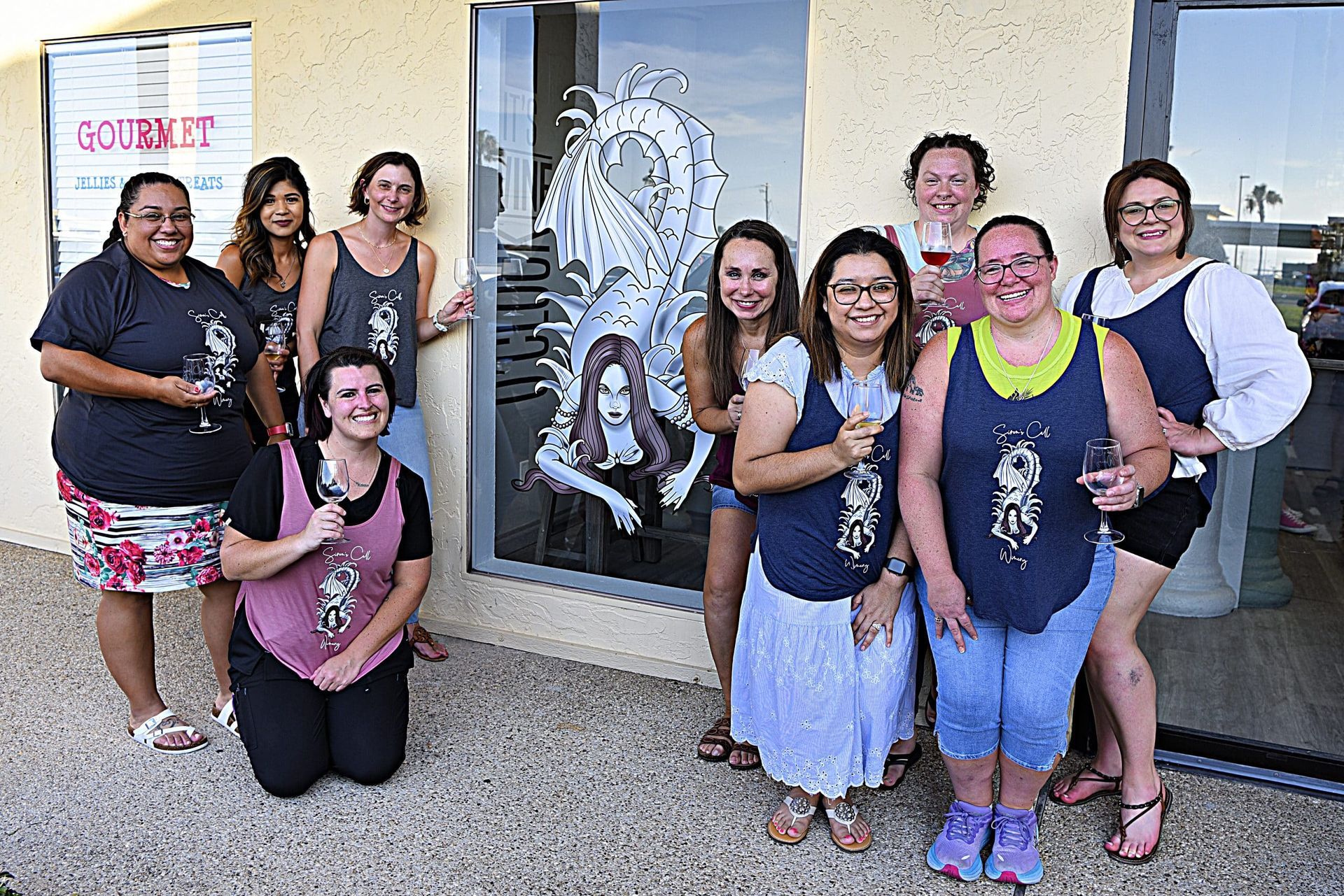 Group of women posing outside a storefront, smiling and kneeling together in front of a window display.