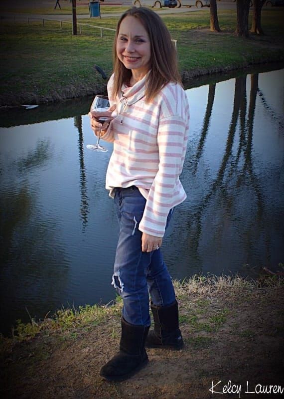 Woman standing by a pond, smiling and holding a drink, wearing a striped top, jeans, and boots.