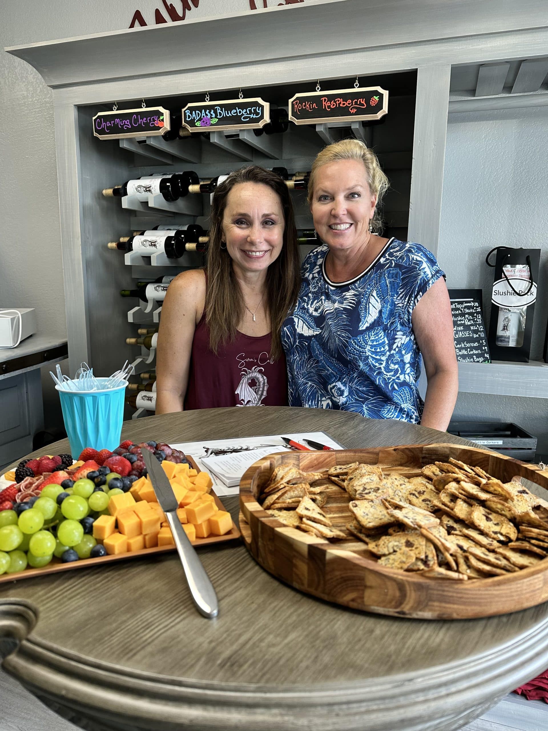 Two women behind a counter with fruit, crackers, and drinks in a kitchen or café setting