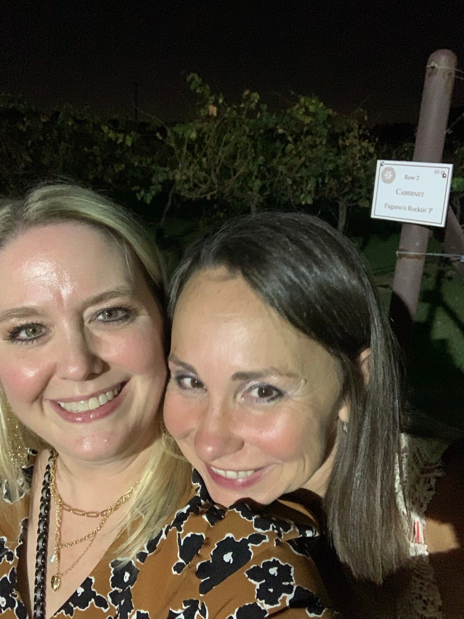 Two smiling people posing outdoors at night, with trees and a sign in the background
