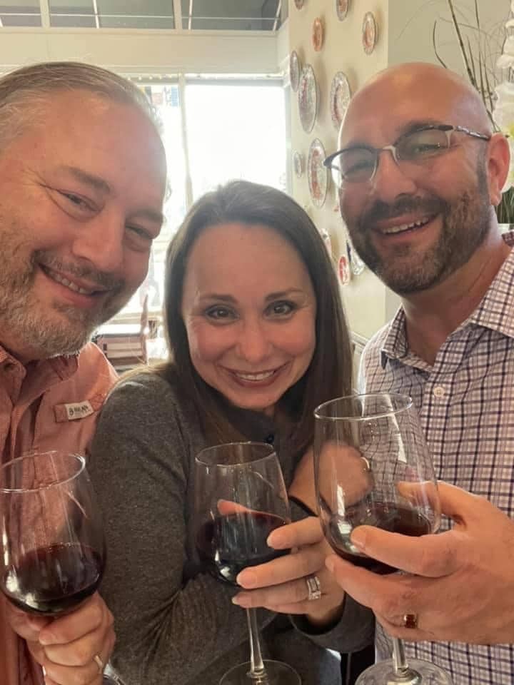 Three smiling people clink wine glasses indoors in a bright restaurant-like setting.