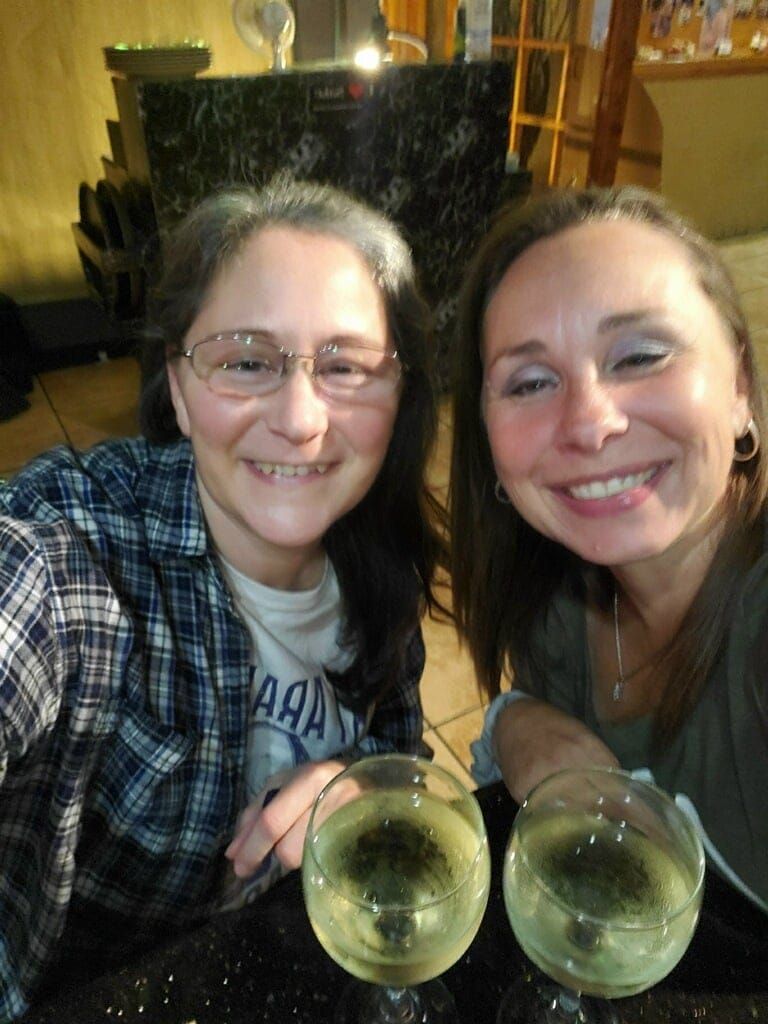 Two smiling friends at a table, holding glasses of white wine in a cozy indoor setting.