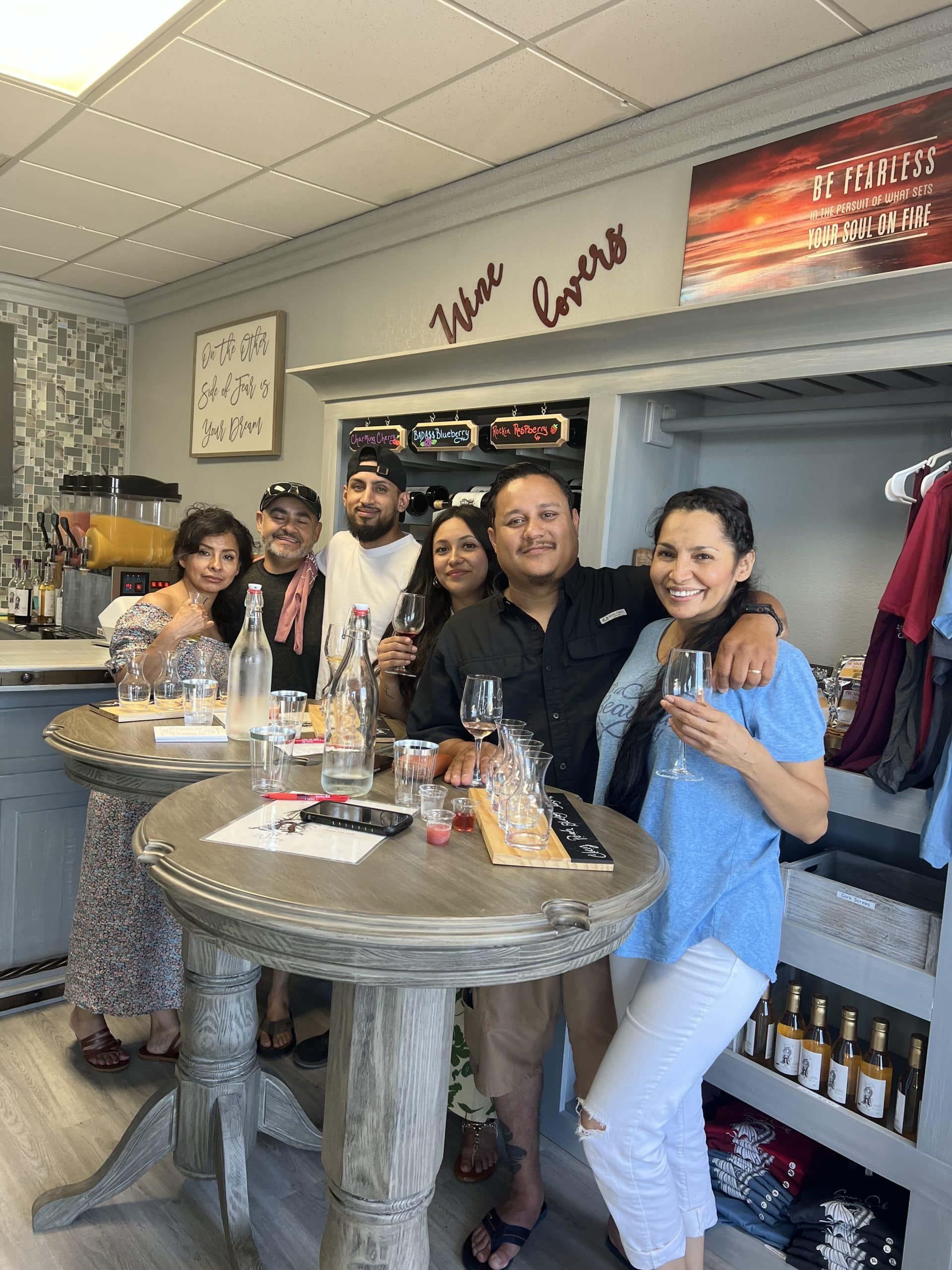 Group of people smiling and holding drinks at a bar counter with shelves of bottles in the background