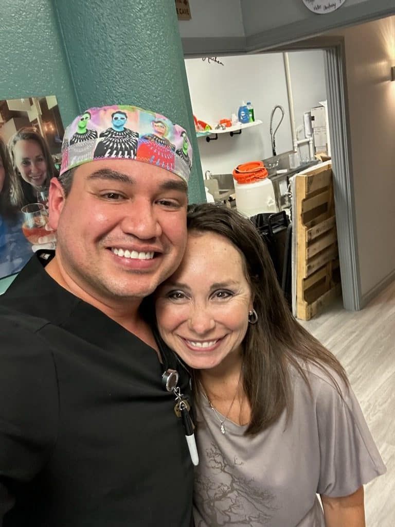 Two people smiling in a medical office, one wearing a colorful scrub cap.