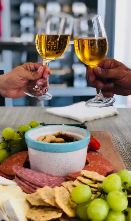 Hands toasting with white wine glasses over a charcuterie board with grapes and crackers