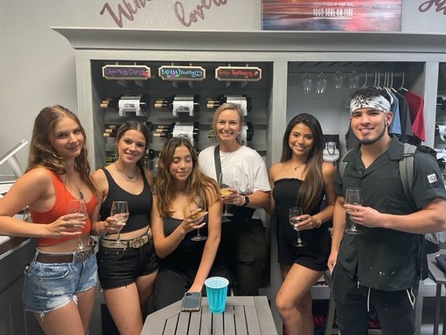Group of six people posing with drinks at a bar, smiling in front of liquor shelves.