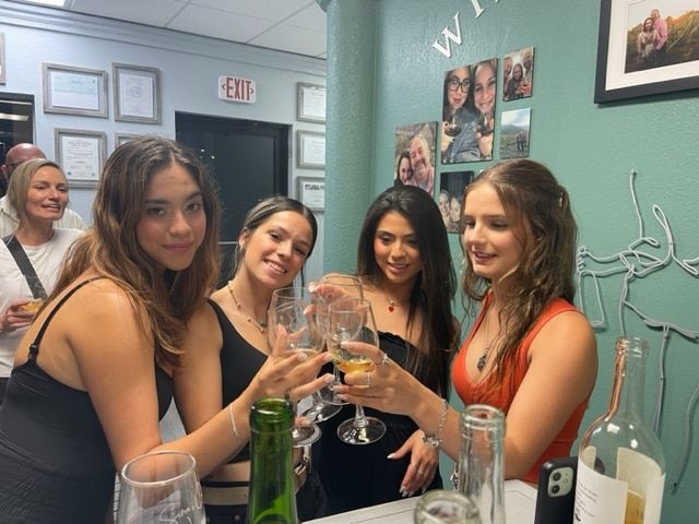 Five women toast with drinks in a teal-walled room decorated with framed photos.