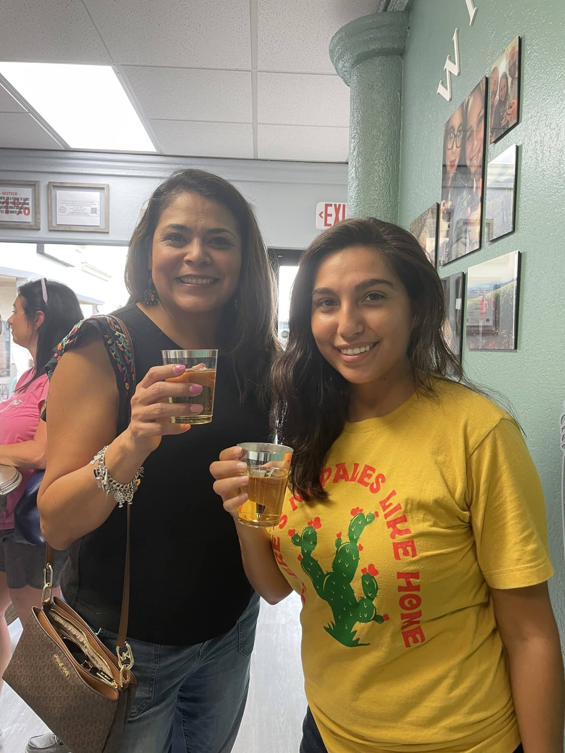 Two women smiling and holding drinks in an indoor event space, one in a black top and one in a yellow shirt.