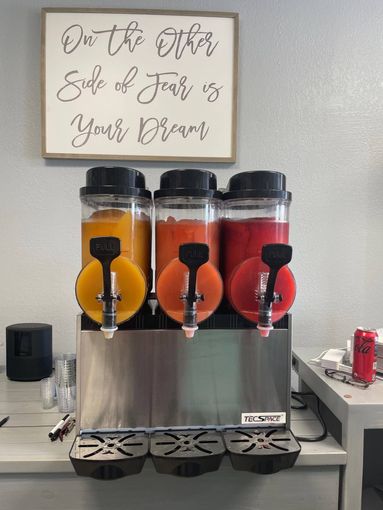 Triple beverage dispenser with orange and red drinks on a counter under a framed sign.