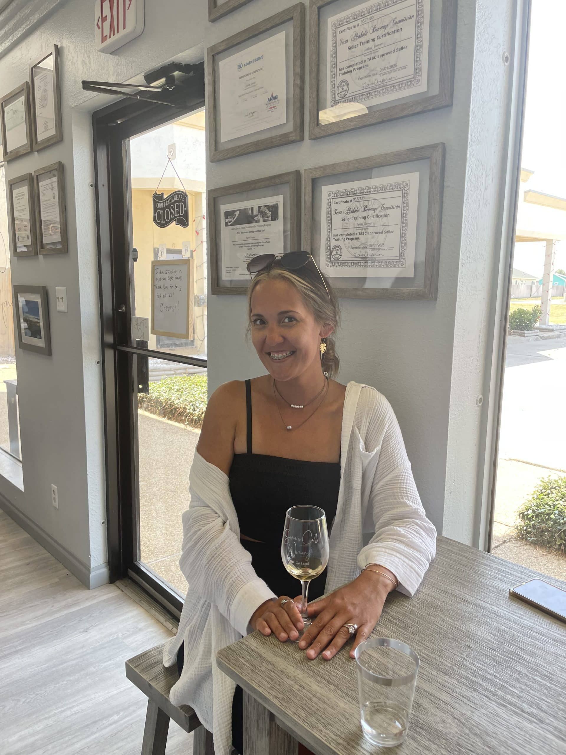 Woman smiling at a table in a bright café, holding a wine glass beside framed certificates.