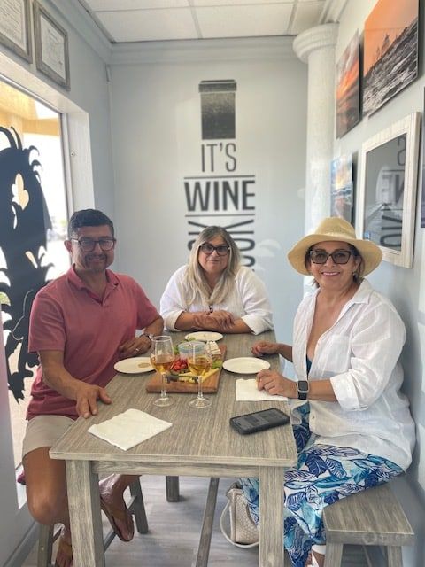 Three people seated at a small table in a bright wine room with snacks and a “It’s Wine Time” sign.