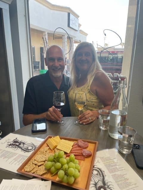 Couple seated at a table outdoors with wine glasses and a plate of grapes and salami
