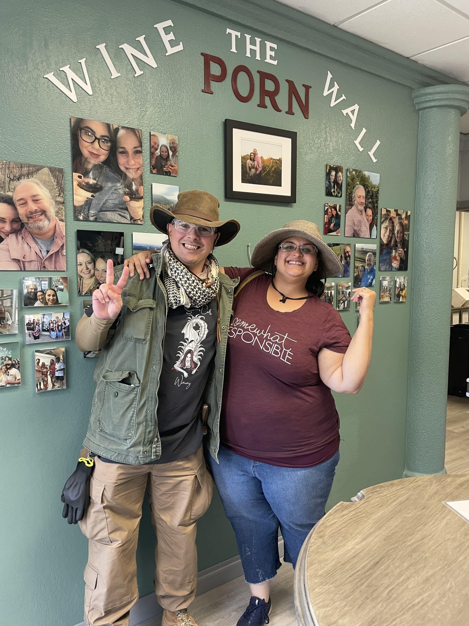 Two people pose smiling in front of a wall of wine bottle labels and photos.