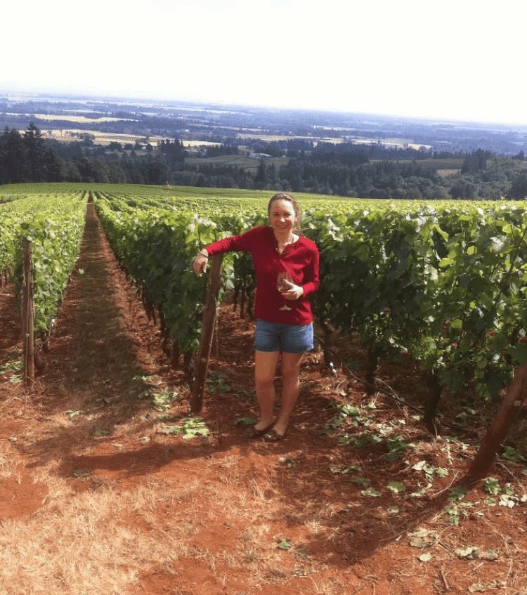 Person standing in a vineyard row, wearing a red top and denim shorts, with fields stretching into the distance.