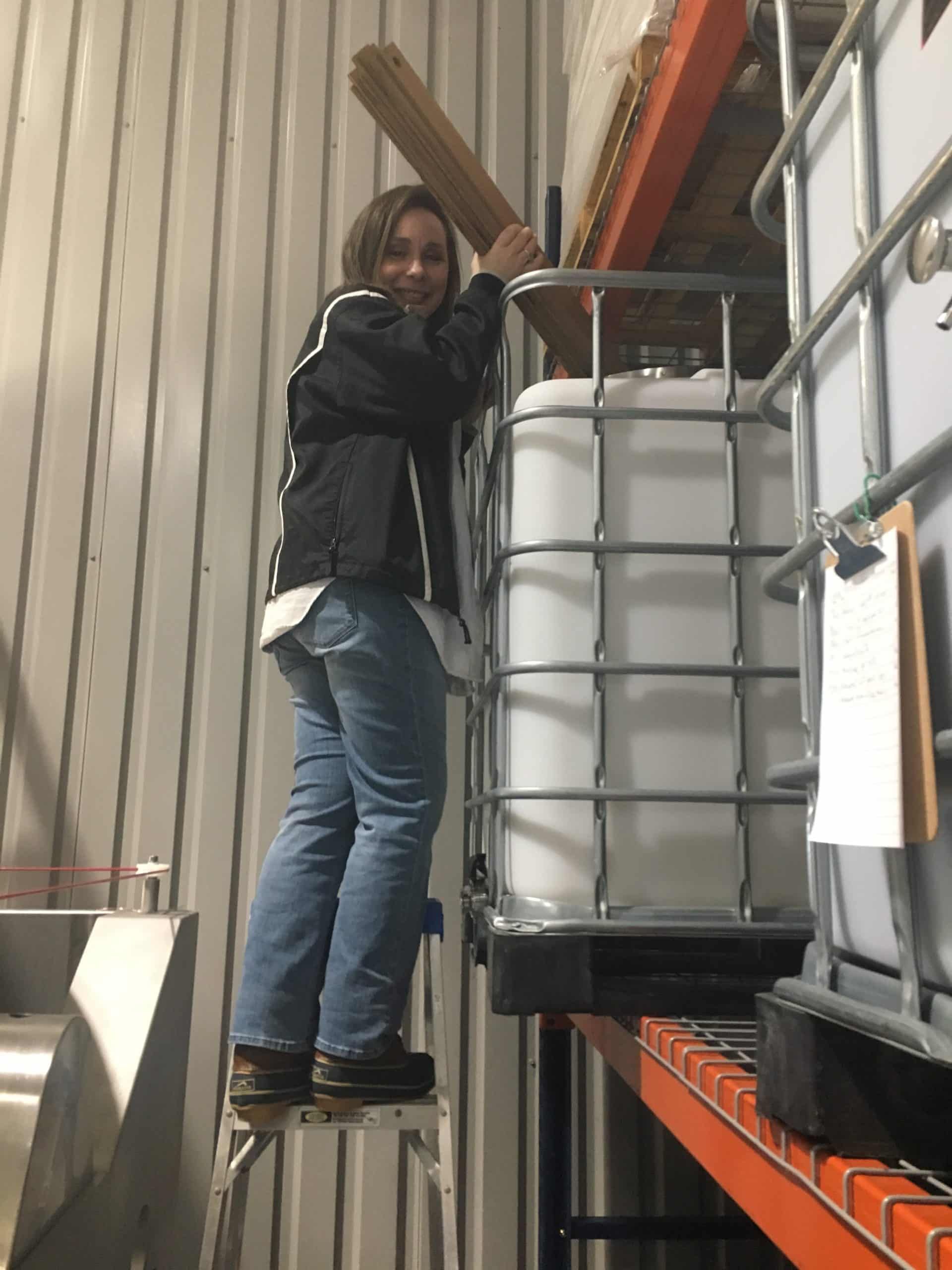Person standing on a stool next to stacked industrial containers in a warehouse, reaching upward.