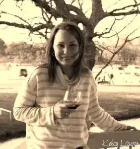 Woman in a striped hoodie smiling outdoors, holding a wine glass near a tree.