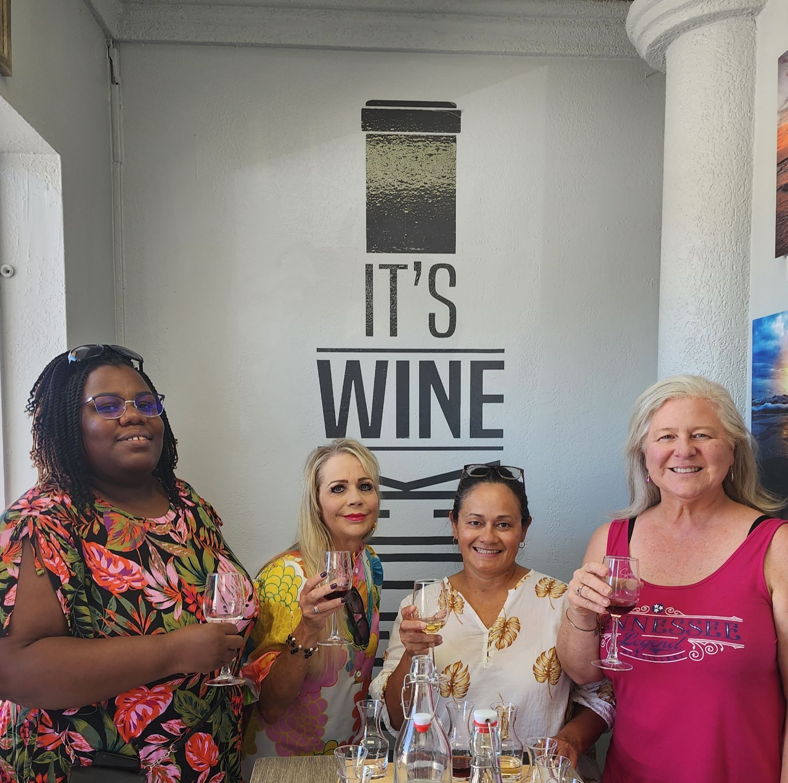 Four women smiling and holding wine glasses under a wall sign that says “IT’S WINE.”