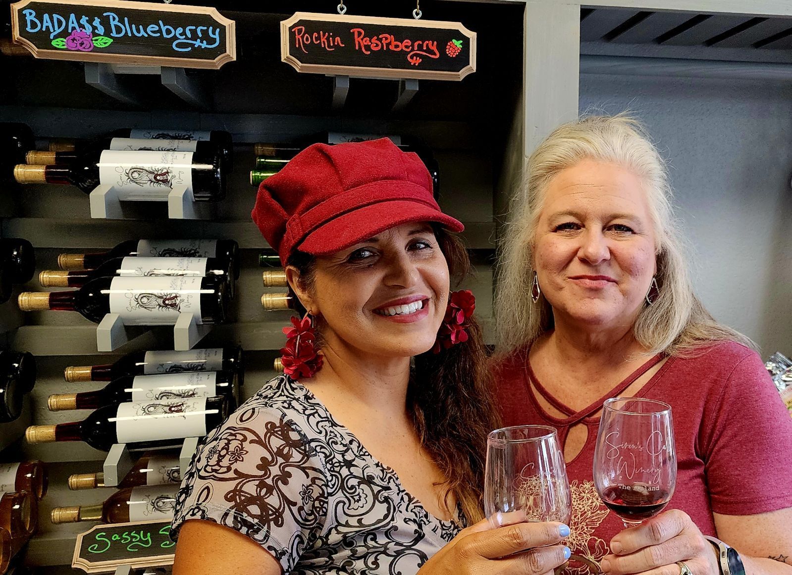 Two women smiling in a shop, holding wine glasses beside a wine rack with labeled bottles