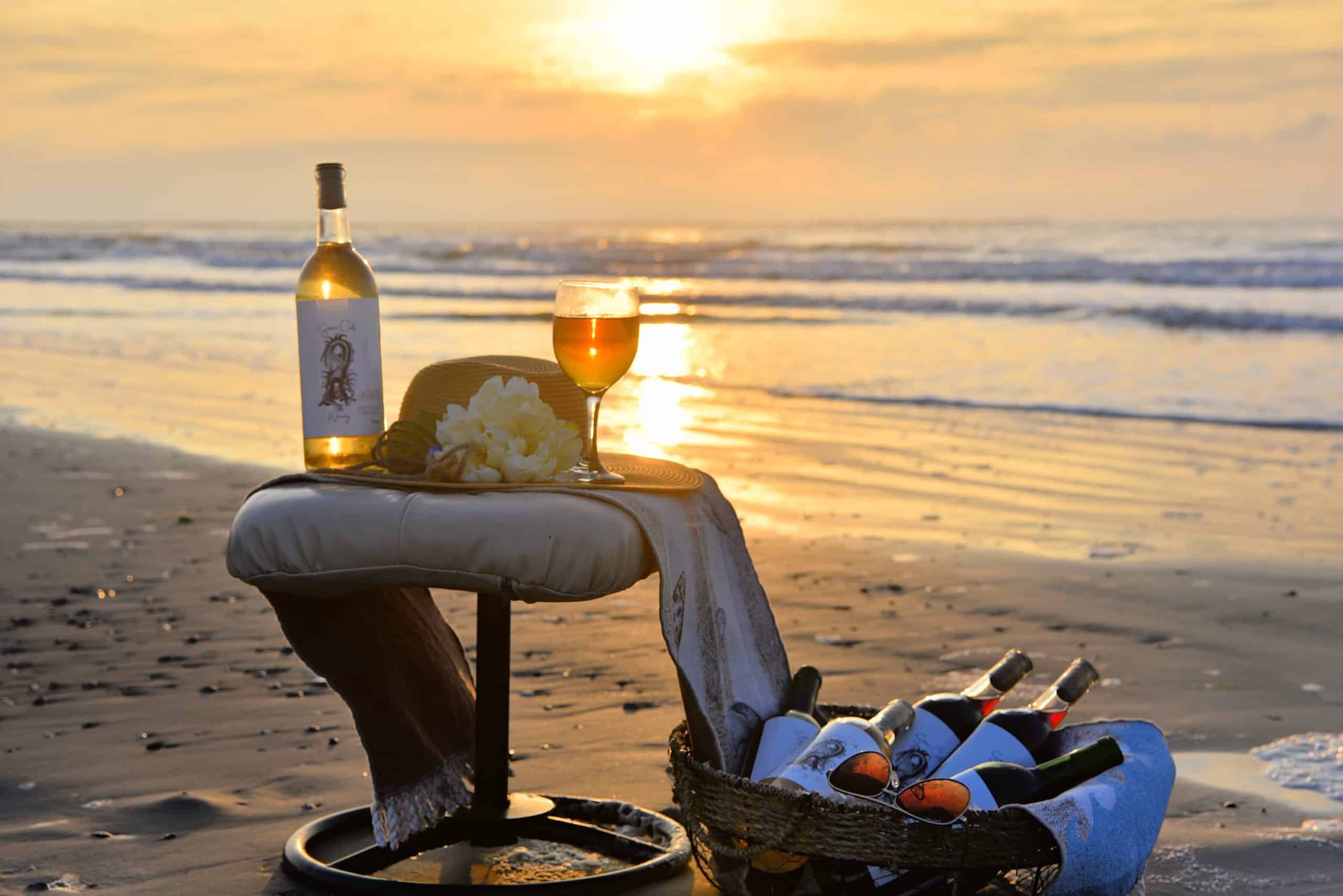 Wine bottle and glass on a beach chair at sunset beside a bucket of ice and bottles on the shore