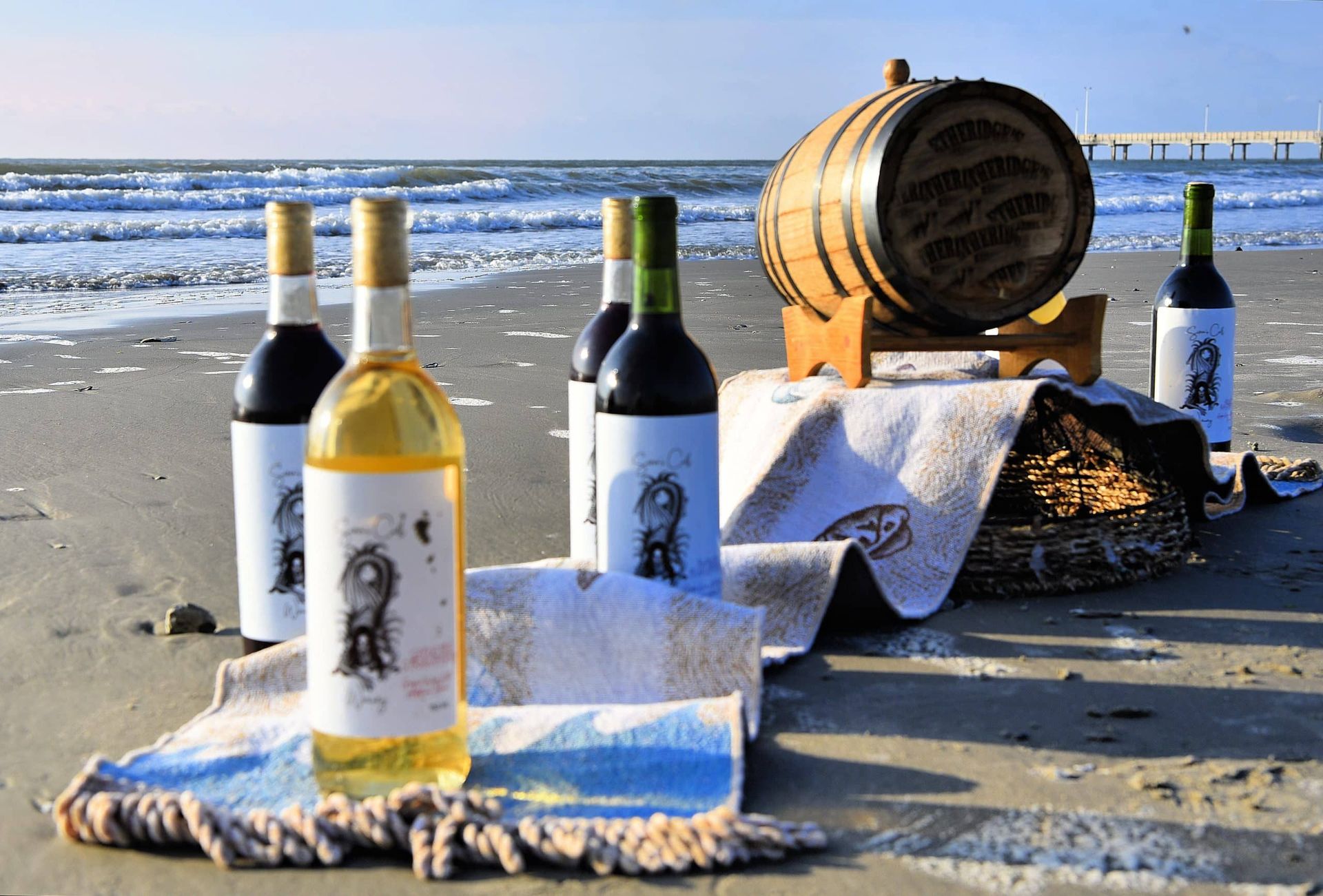 Wine bottles and glasses on a beach table beside a barrel, with ocean waves in the background