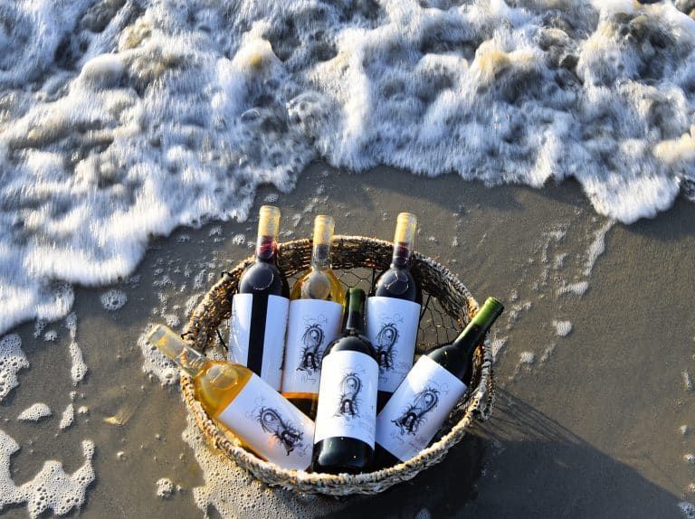 A basket of wine bottles on a beach near foamy surf at the water’s edge