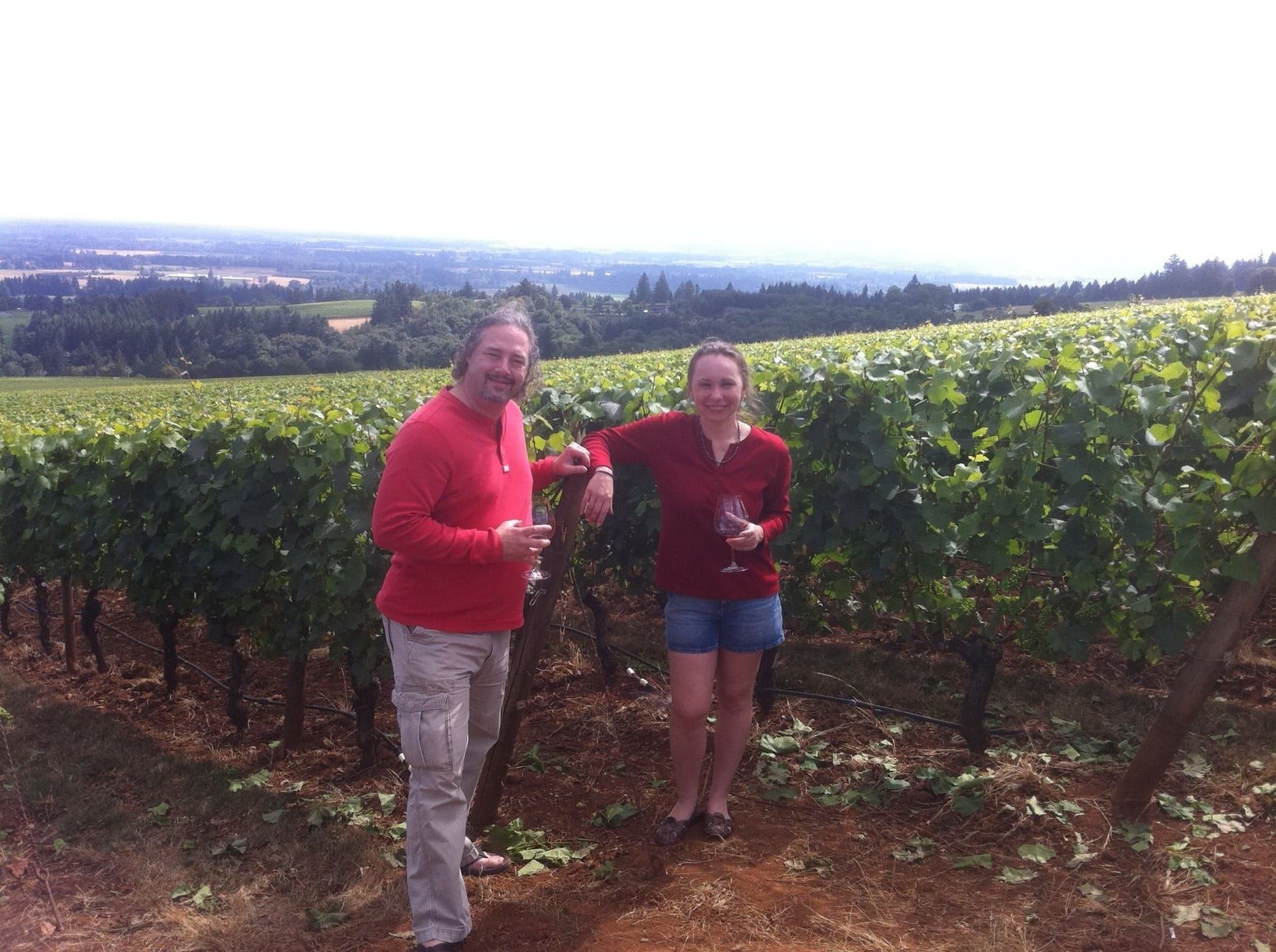 Two people standing in a vineyard holding wine glasses, with rolling hills in the background.