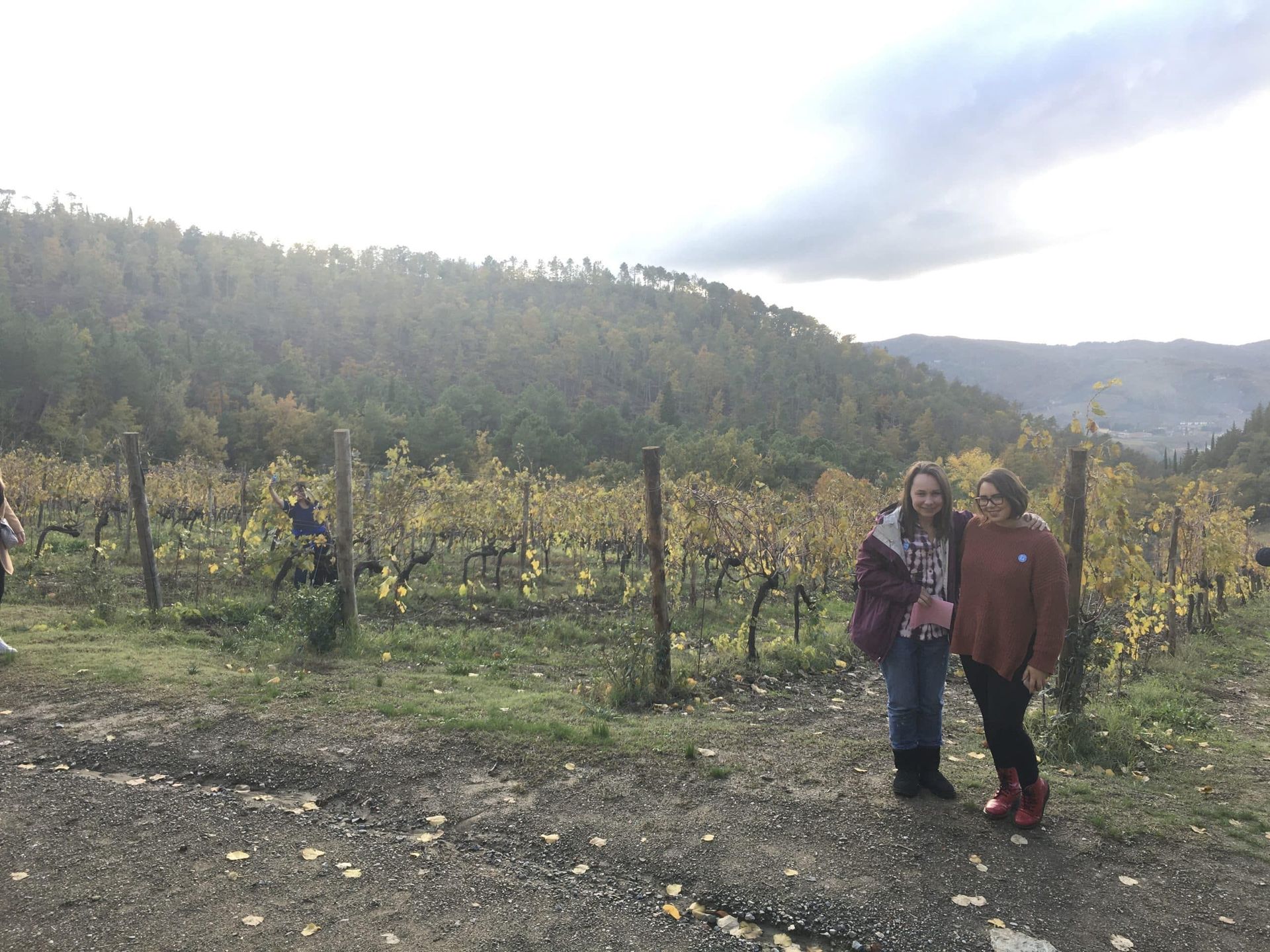 Two people standing in a vineyard with hills and cloudy sky in the background.