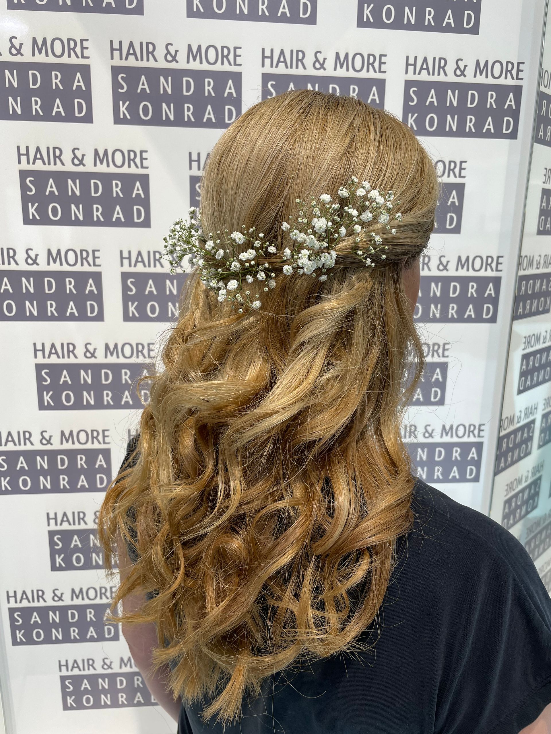 Frau mit welligem blonden Haar, geschmückt mit einem Blumenkranz, vor einer Salonkulisse.