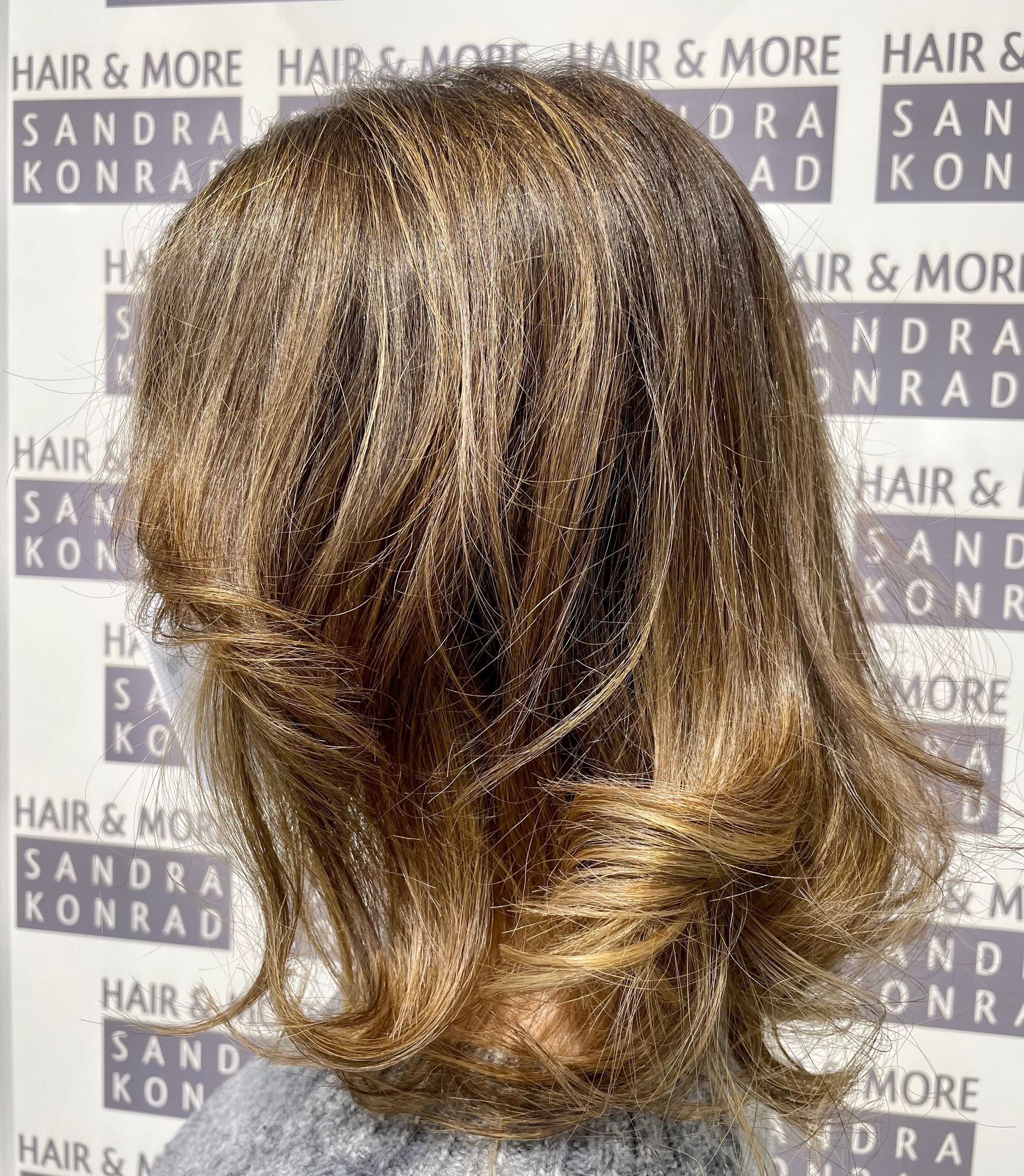 Schulterlanges, gestuftes braun-blondes Haar, möglicherweise gestylt. Im Hintergrund ist „Hair & More Sandra Konrad“ zu sehen.