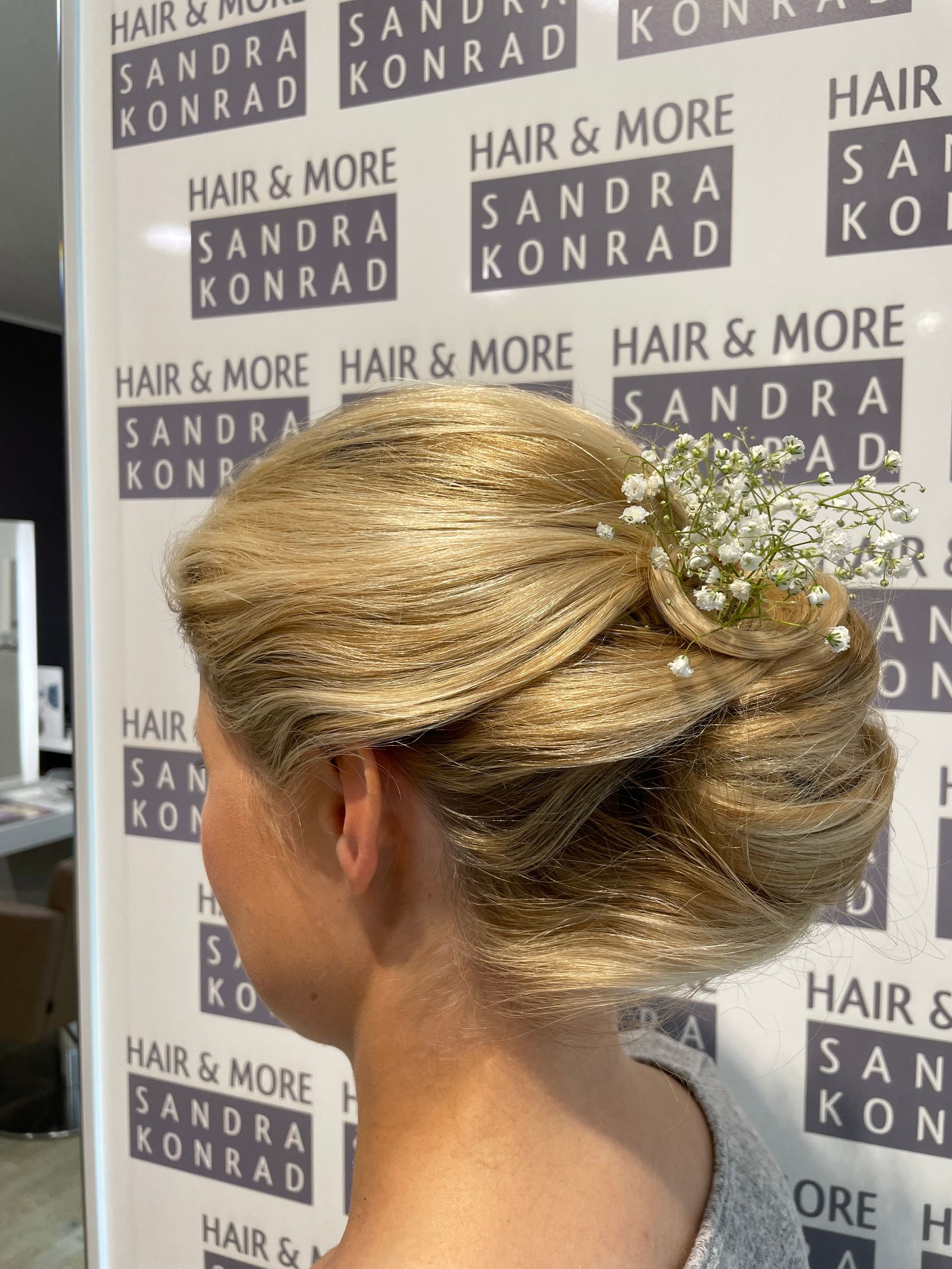 Frau mit blonder Hochsteckfrisur, verziert mit Schleierkraut, vor einer Salonkulisse.
