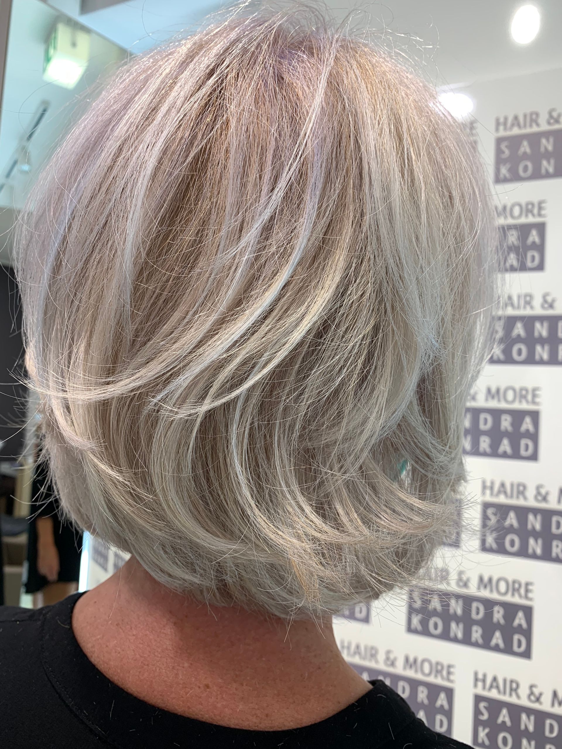 Rückenansicht einer Frau mit kurzem, gestuftem, silberblondem Haar. Salon-Hintergrund mit Text.
