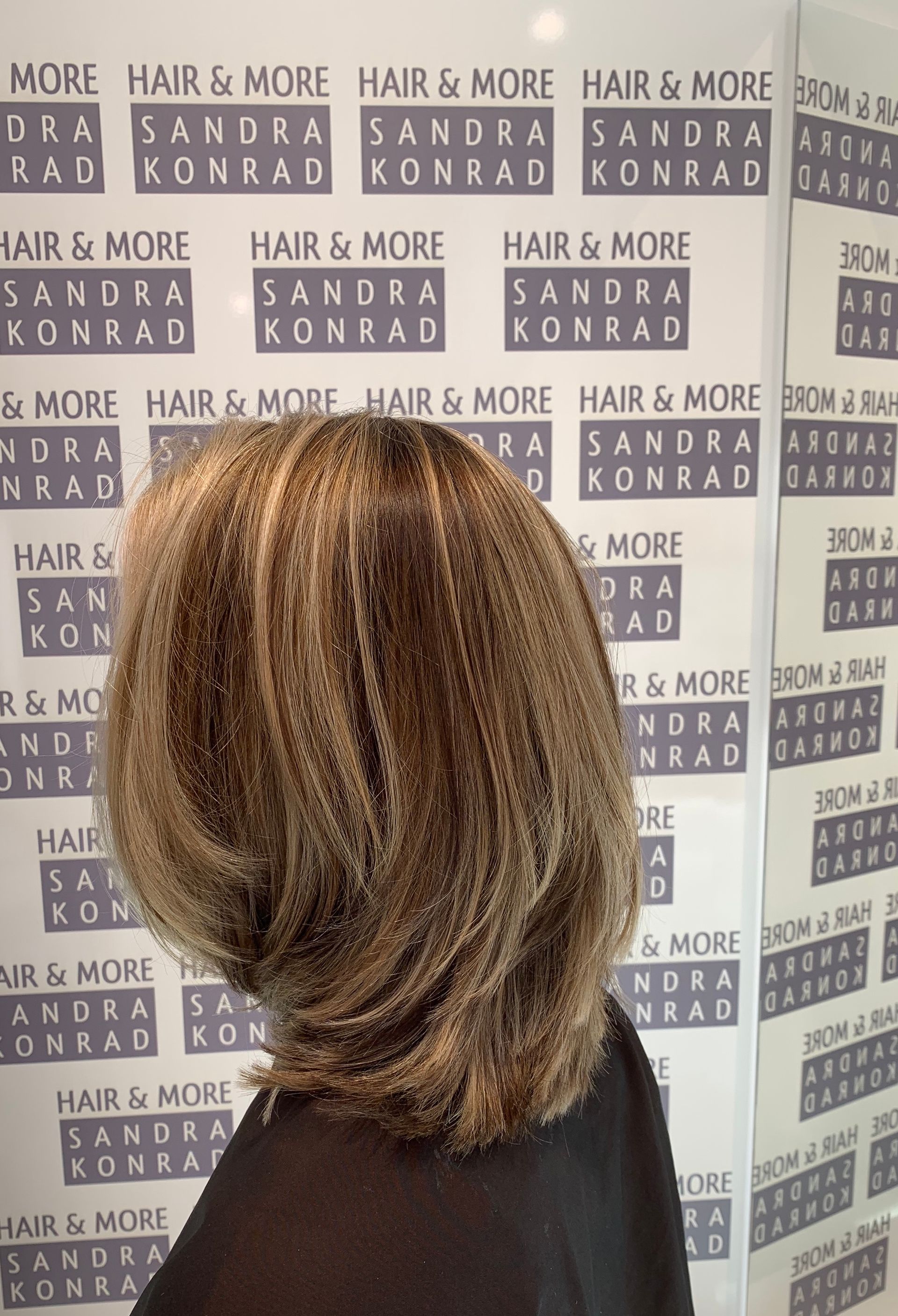 Frau mit gestuftem, braunem Haar vor einer Salonwand mit dem wiederholt aufgedruckten Schriftzug „Hair & More Sandra Konrad“.