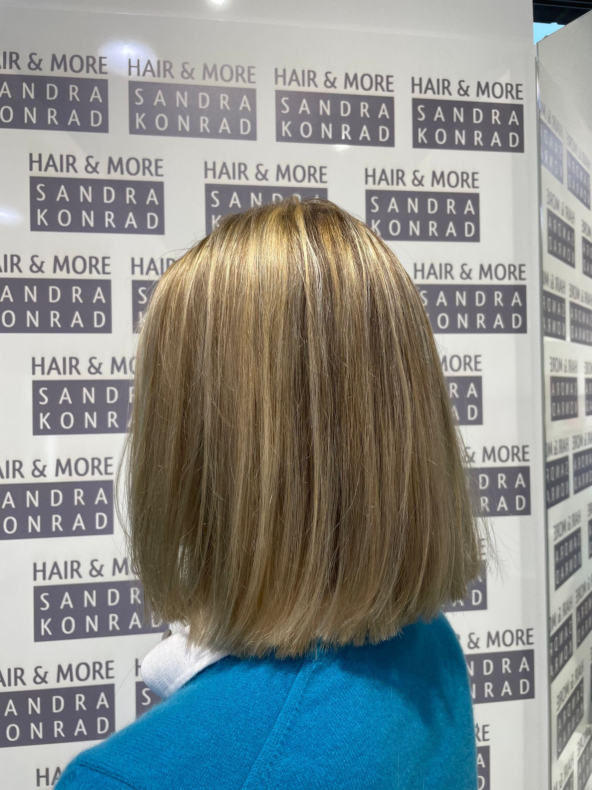 Frau mit gekräuseltem, blondem Haar und blauem Pullover, die vor der Wand „Hair & More Sandra Konrad“ steht.