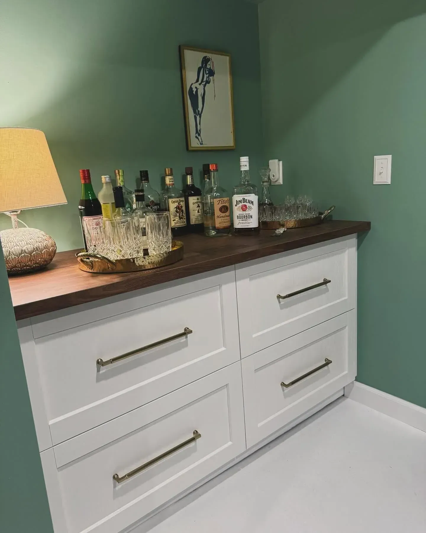 White cabinet with wood top, liquor bottles, and a lamp against sage green walls in a room corner