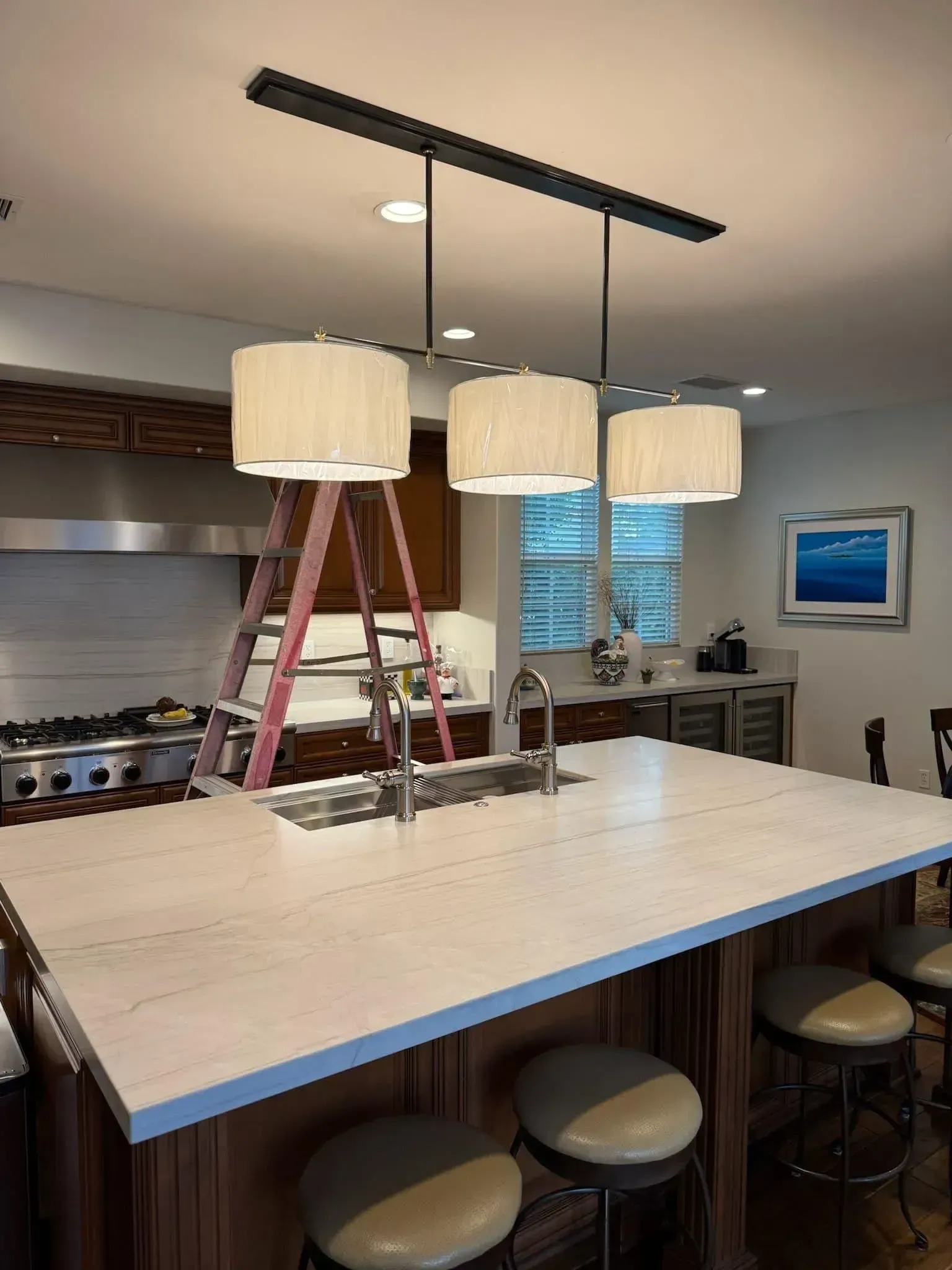 Kitchen island with three pendant lights being installed, ladders beneath a black track over a white countertop.