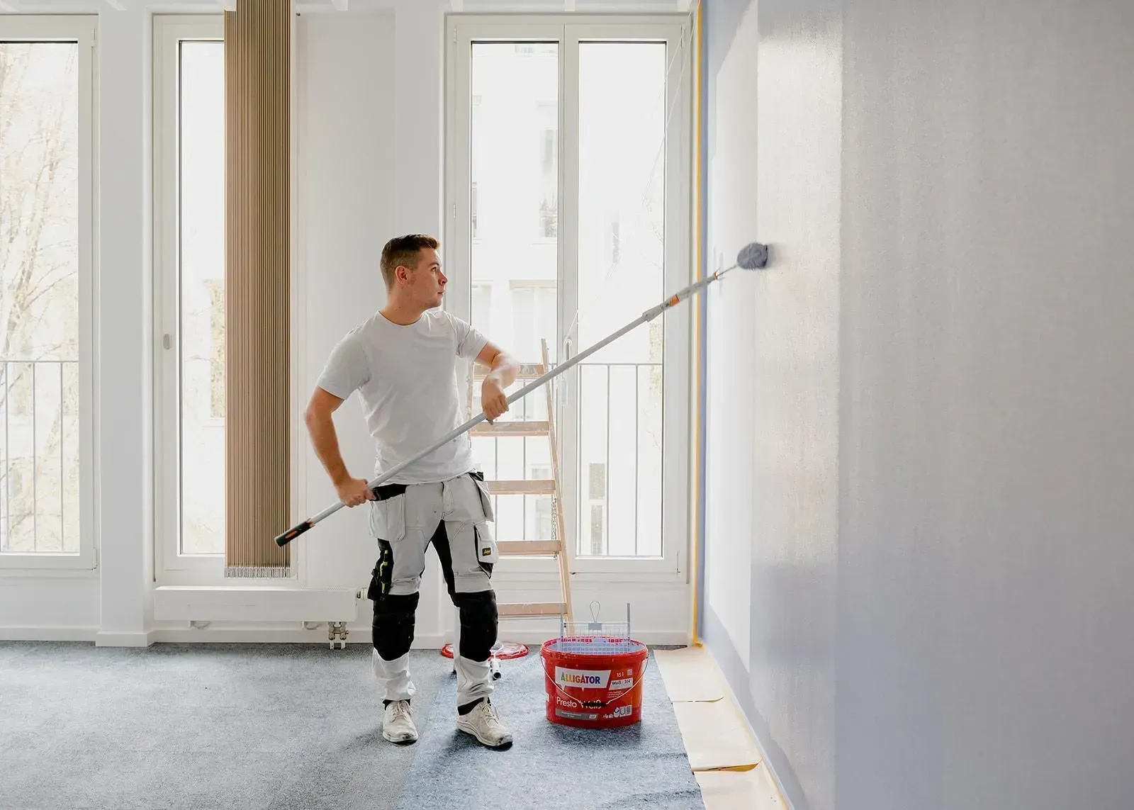Painter rolling white paint on an interior wall in a bright room with large windows