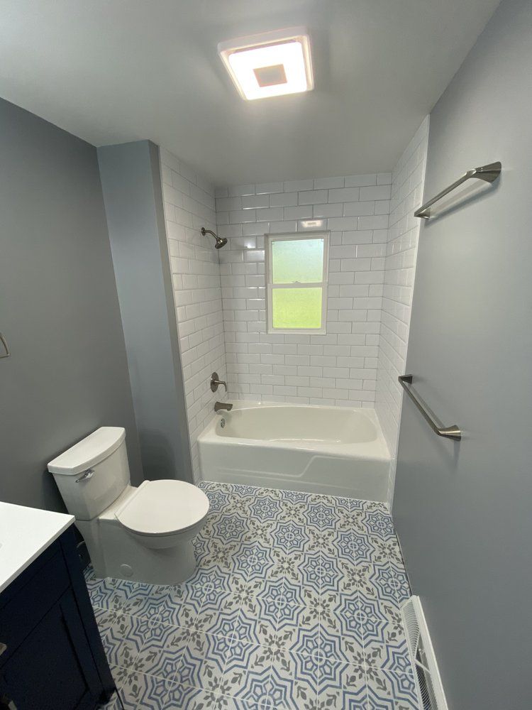 Small bathroom with gray walls, white subway tile tub surround, blue patterned floor, and toilet beside the tub