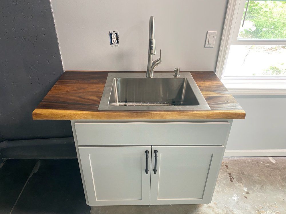 Small kitchen sink cabinet with wood countertop, stainless steel sink, and white doors by a window.