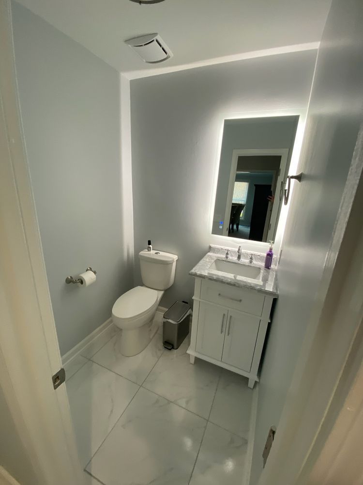 Small bathroom with toilet, vanity, mirror, and gray tile floor, viewed through an open door