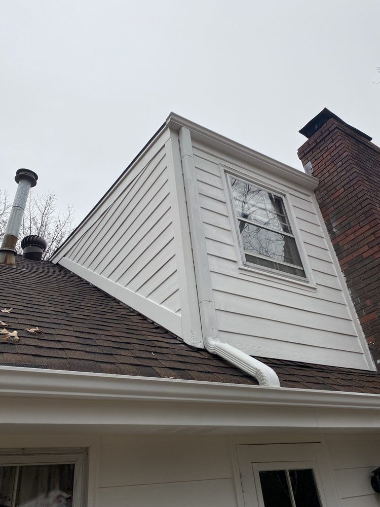 White dormer on sloped shingle roof beside red brick chimney and window under gray sky