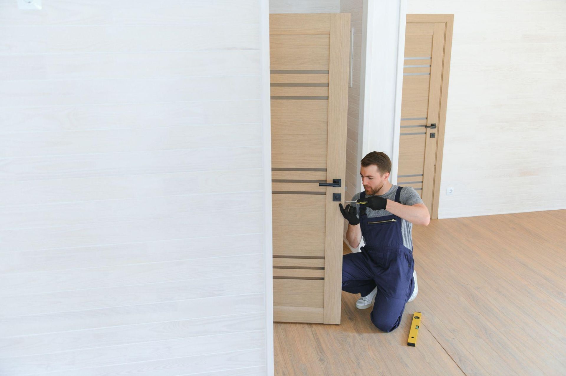 Person kneeling by a wooden door, using a drill in a bright empty room with a tape measure nearby