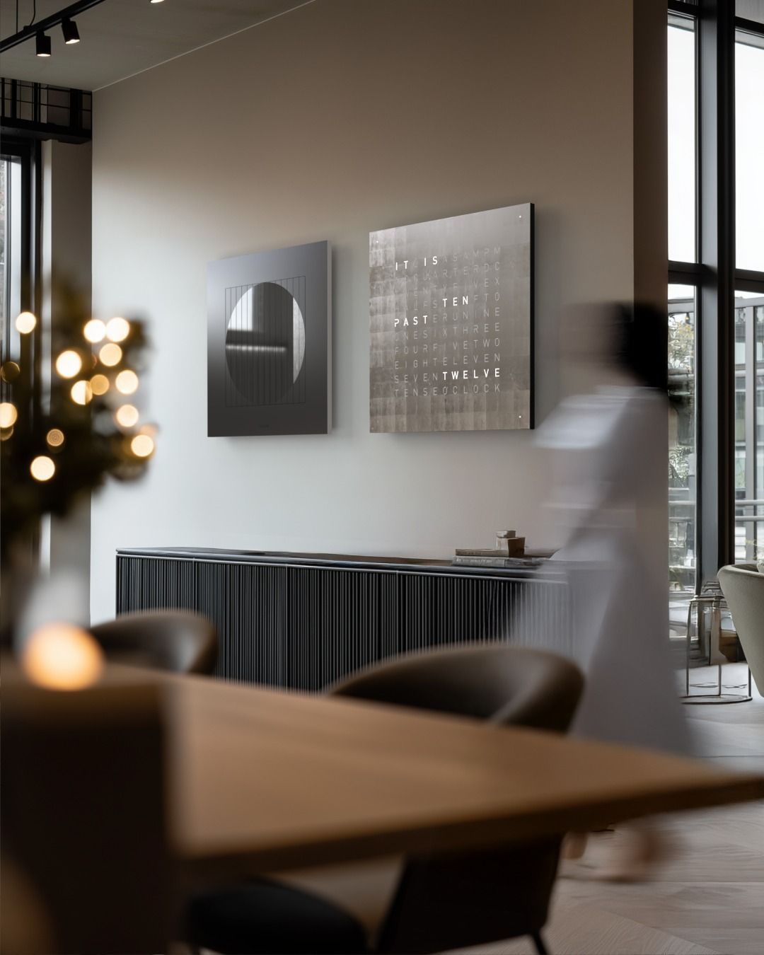 Een ruimte met twee vierkante kunstwerken aan de muur boven een donker dressoir met latten, naast een bewegend persoon.
