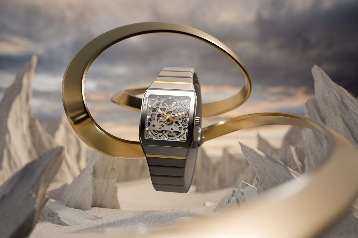 Gouden horloge met een metalen band, tentoongesteld tussen wervelende gouden ringen tegen een rotsachtige achtergrond.