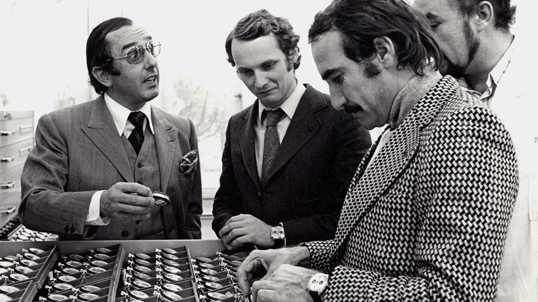 Drie mannen in bespreken spullen die op tafel liggen uitgestald; een van hen gebaart, terwijl de ander naar beneden kijkt.