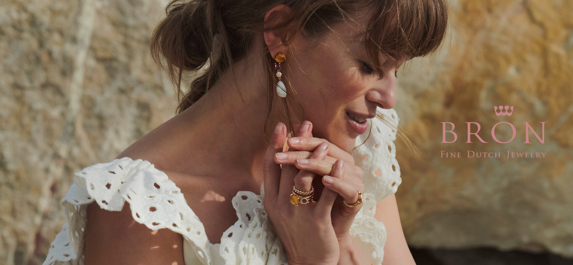 Een vrouw in een witte top draagt ​​gouden oorbellen en ringen, haar handen gevouwen bij haar gezicht, bij een rotswand.