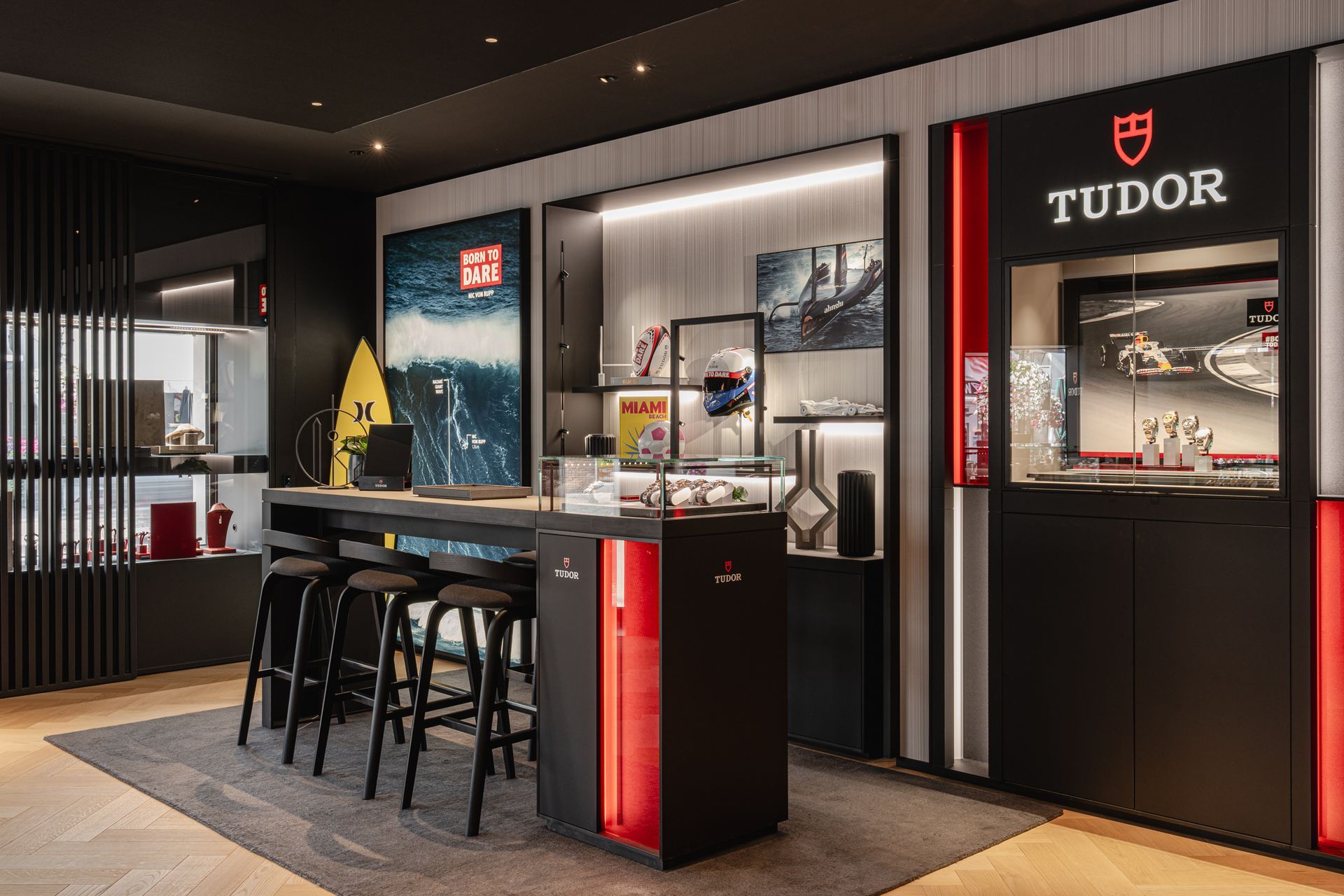 Interieur van een luxe horlogeboetiek met Tudor-logo, vitrines en een zwarte bar met barkrukken.