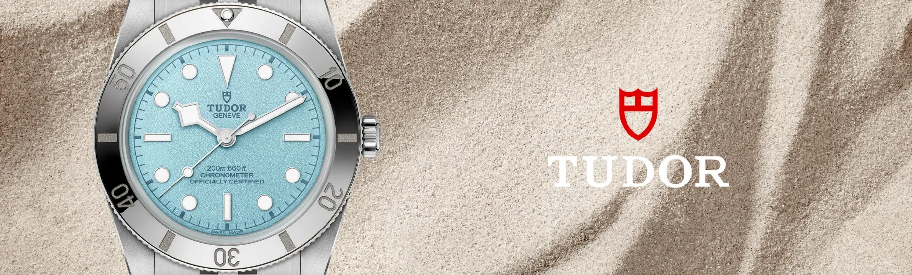 Tudor polshorloge met een turquoise wijzerplaat en zilveren band op een zandkleurige achtergrond, met rood Tudor-logo.