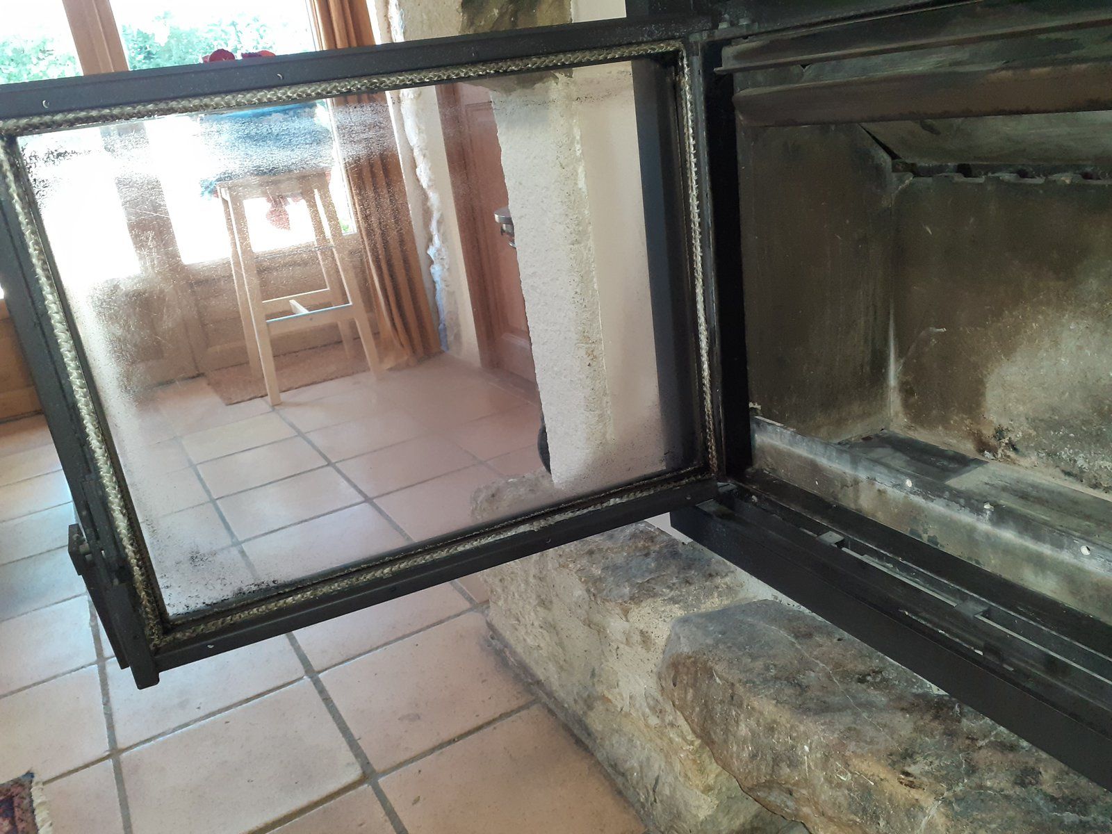 Cheminée ouverte avec porte vitrée, laissant apparaître une pièce carrelée se reflétant à l'intérieur et un foyer en pierre en dessous.