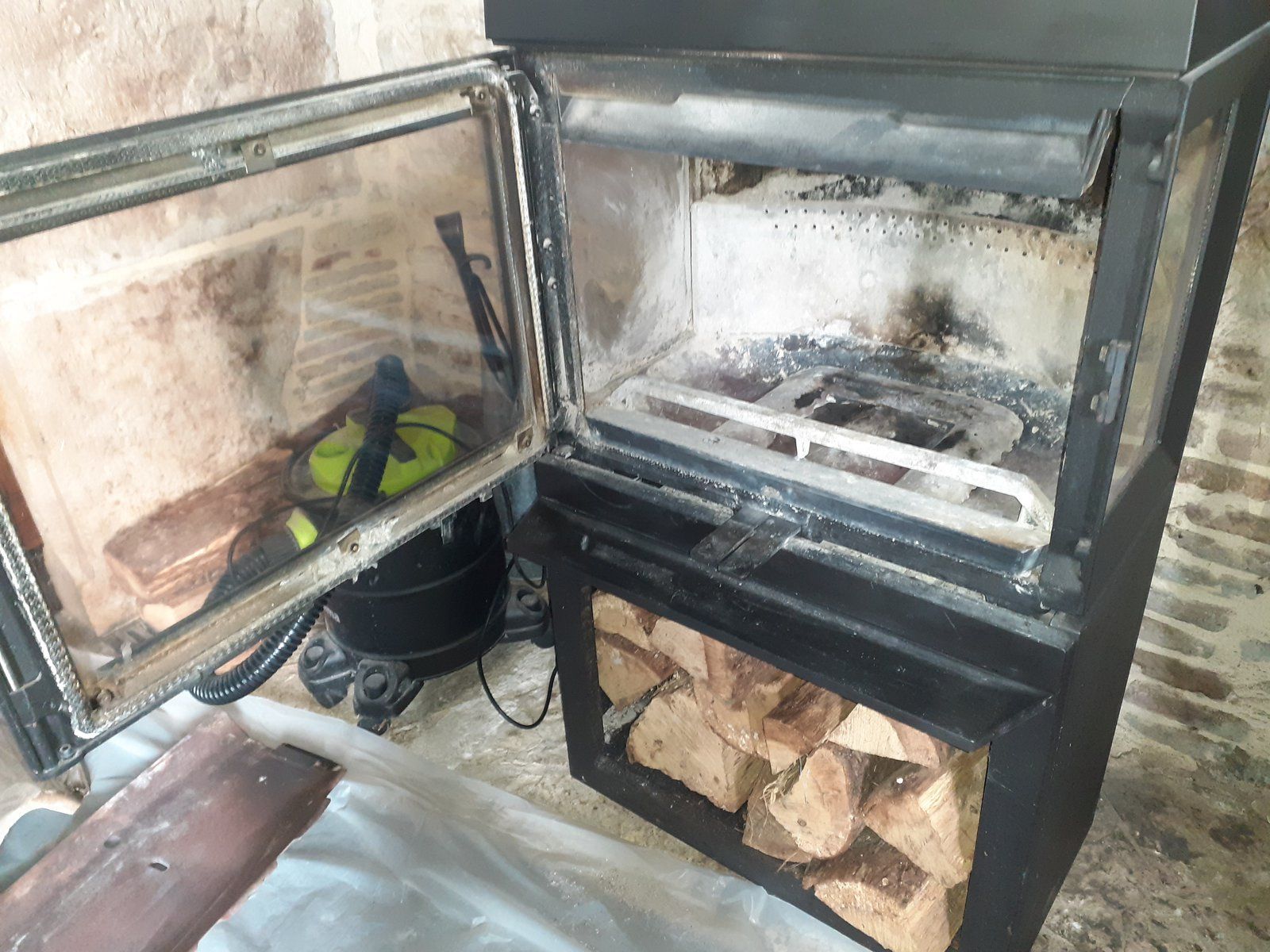 Poêle à bois noir avec porte ouverte, bois de chauffage stocké en dessous et étagère murale à côté.